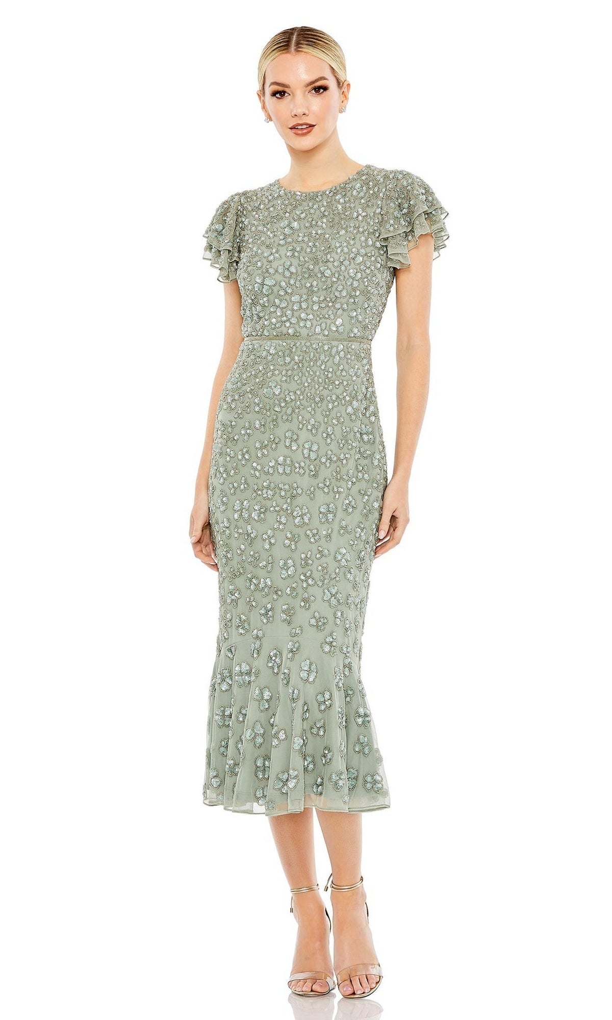 A woman wears the sage green Short Wedding Guest Dress 10771 by Mac Duggal, featuring a fitted midi silhouette, short ruffled sleeves, beaded details, and a flounced hem. She pairs it with silver heels for a chic semi-formal look.